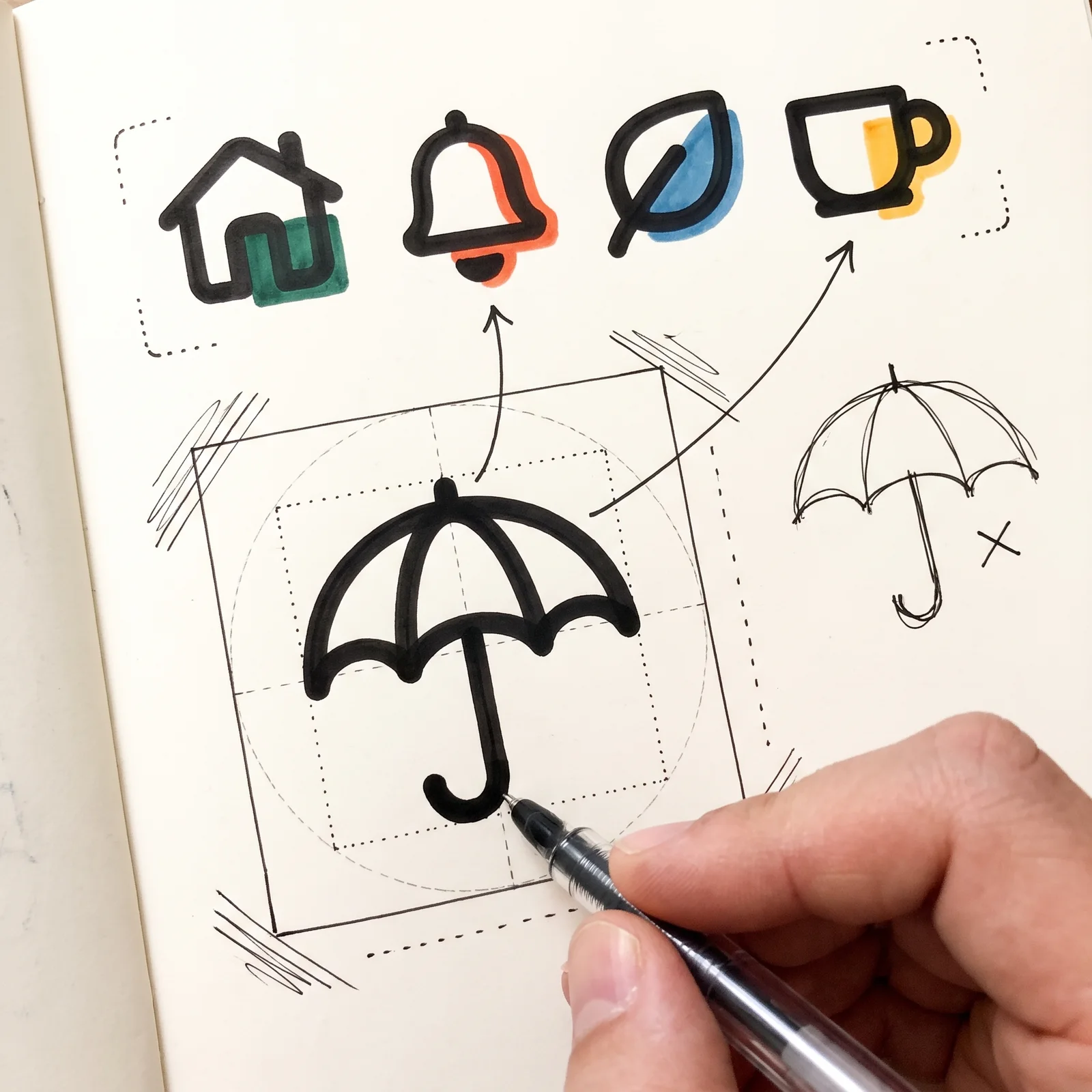 A hand draws an umbrella icon on a square keyline grid below a row of matching icons of a house, bell, leaf and cup, while a thin freehand umbrella sits aside with a cross.