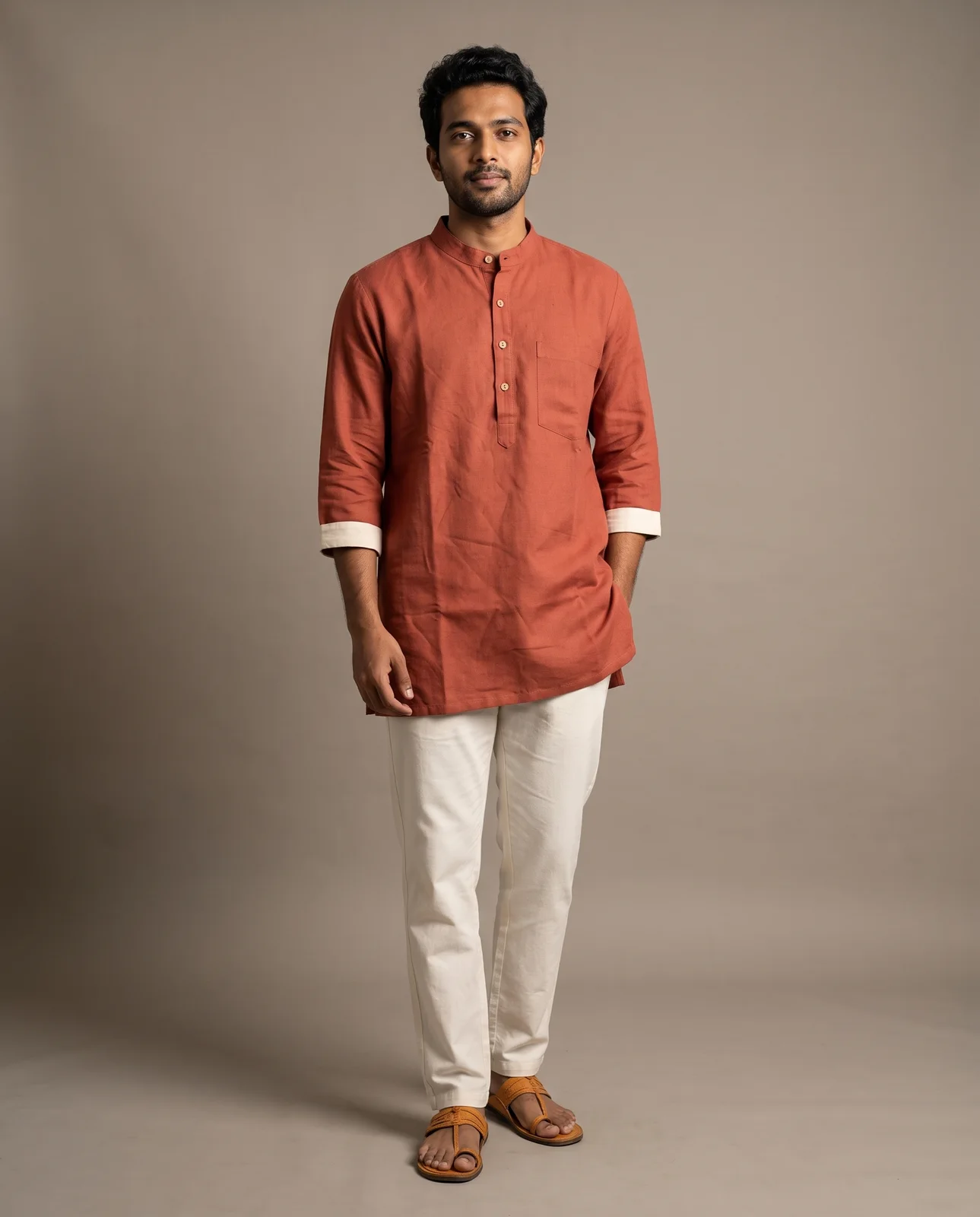 A man in his late twenties on grey paper wearing the rust kurta with off-white trousers and tan sandals, looking into the lens.