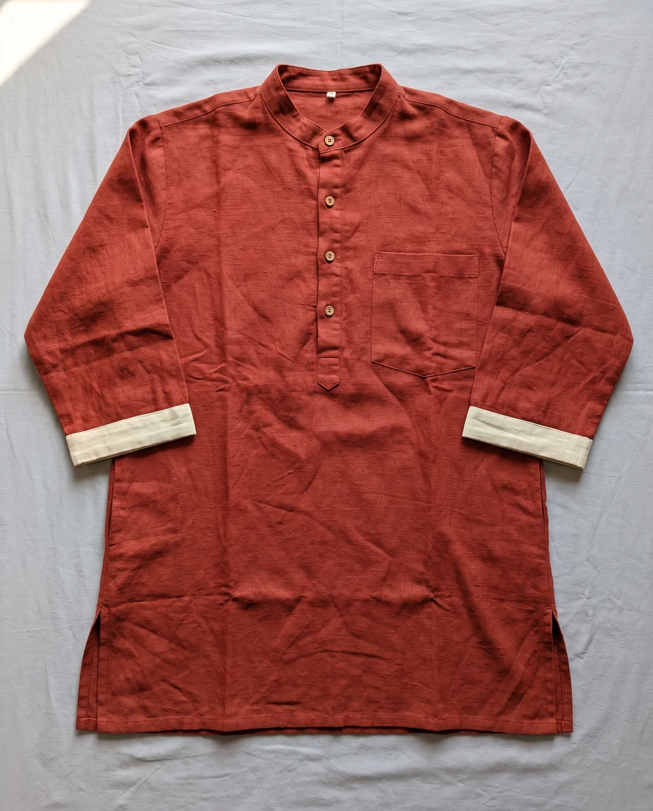 Phone photo from above of a rust linen short kurta laid flat on a grey sheet, four wooden buttons, cream cuffs, one chest pocket.