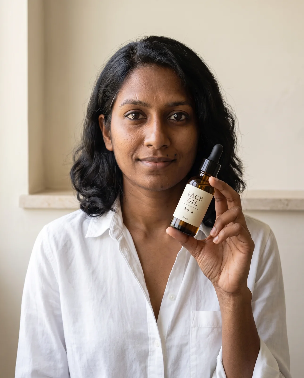 The same woman in a white shirt holding a small amber face oil bottle at collarbone height, label to the camera.
