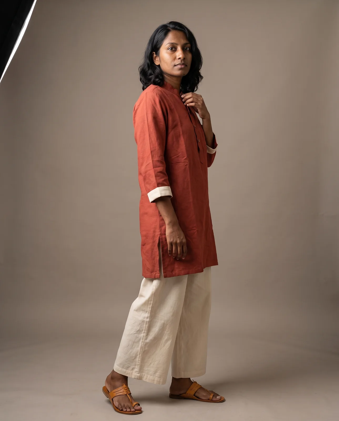 The same woman turned three-quarters in the rust kurta, face back to the lens, a hand at the collar, full length on grey paper.