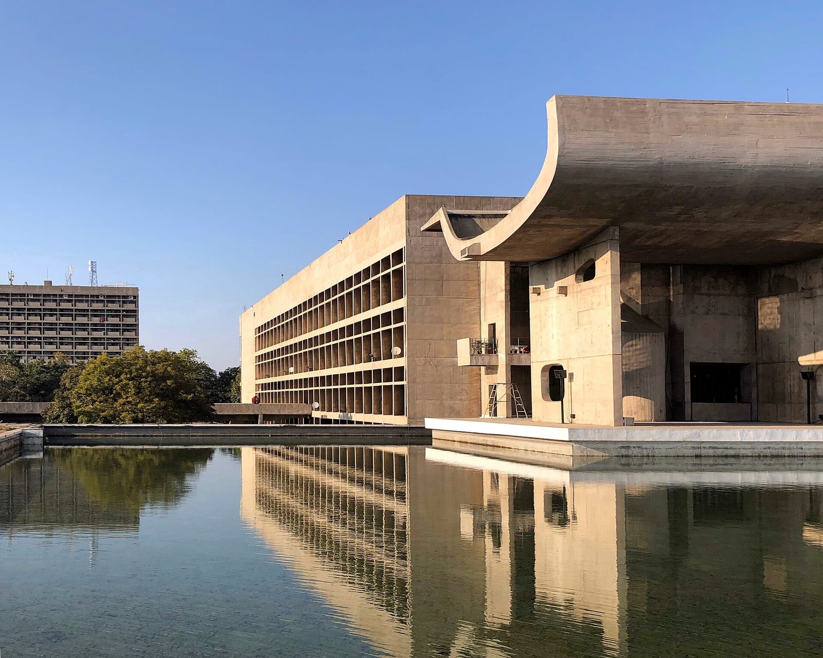 Le Corbusier's Palace of Assembly at Chandigarh photographed across its reflecting pool in low sun: a deep concrete portico under an upturned curved roof, a grid of shaded window bays running down the long side, and the whole building doubled in still water beneath a clear blue sky.